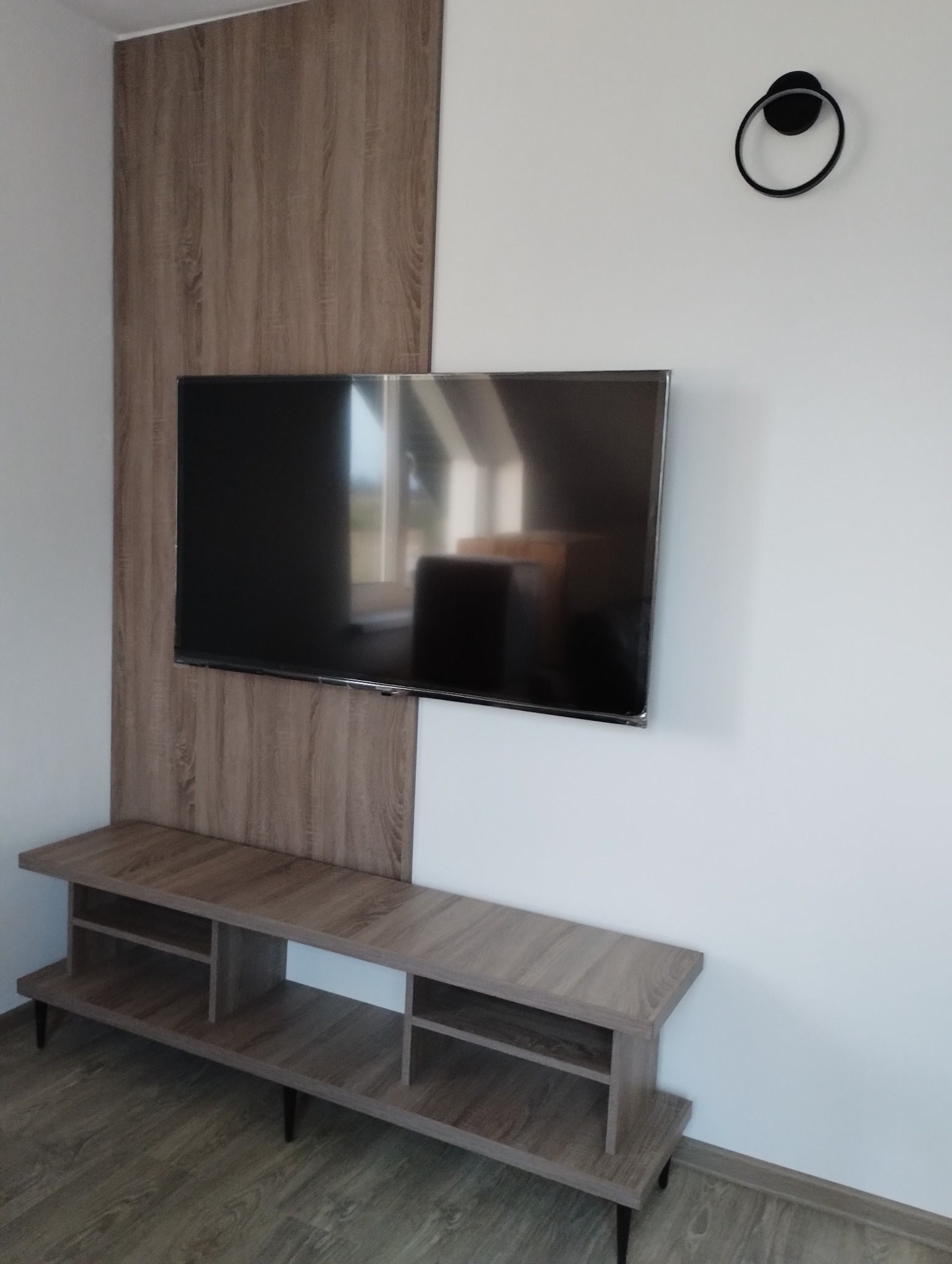 Nowoczesna szafka RTV z drewnopodobną okleiną, zamontowana na ścianie z panelem dekoracyjnym o identycznym wykończeniu, podwieszony telewizor i minimalistyczna lampa ścienna.