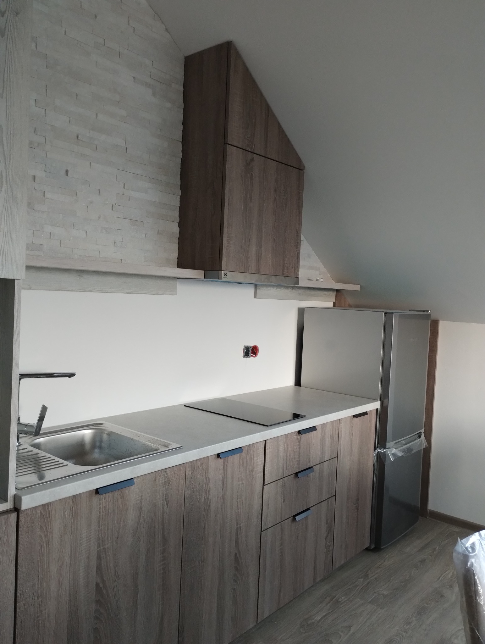 Nowoczesna kuchnia w skosach: szare szafki z drewnianym wzorem, biały blat, ściana z dekoracyjnej cegły, srebrna lodówka i czarna płyta indukcyjna.