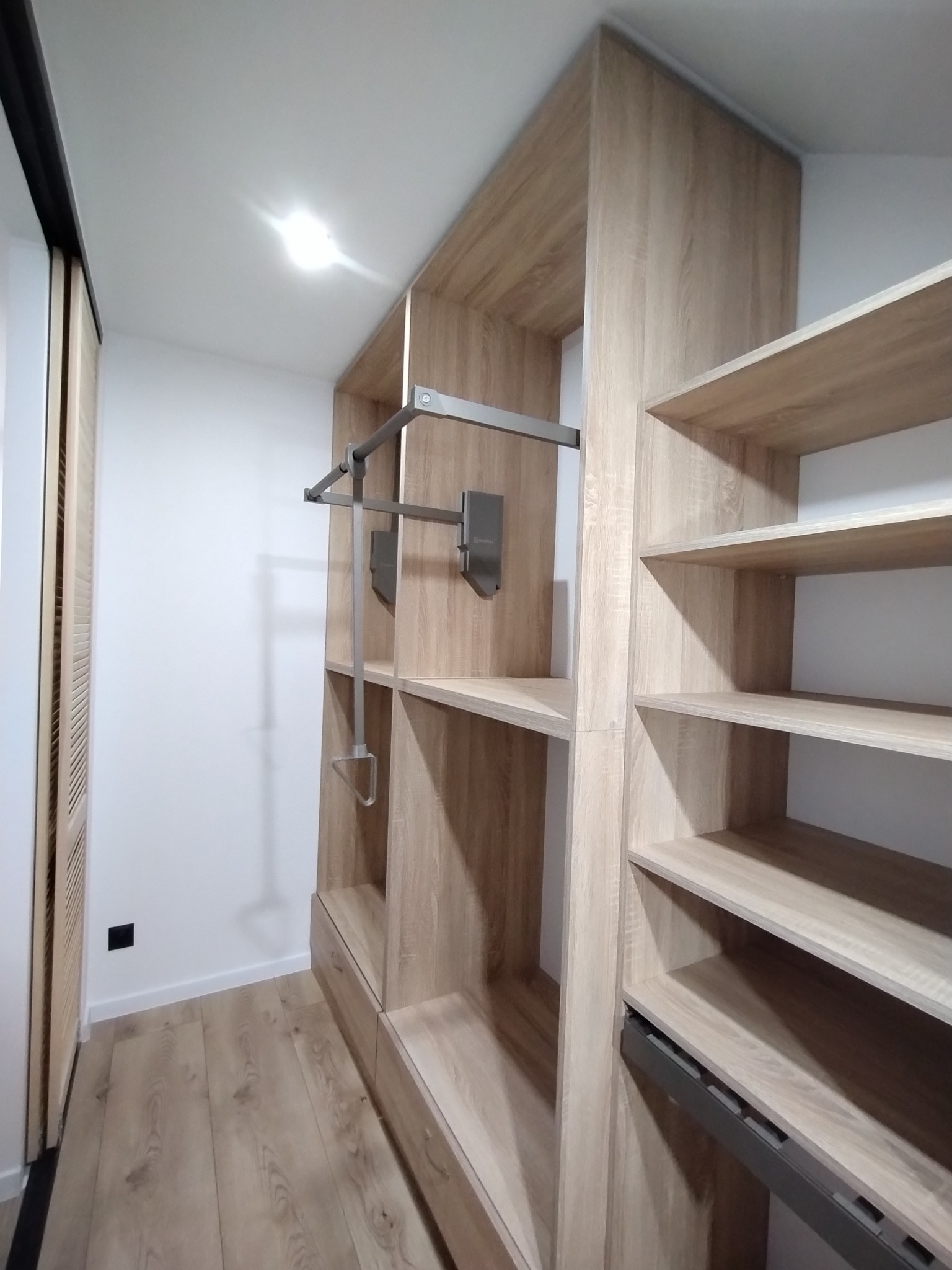 Wnętrze garderoby z jasnego drewna z systemem półek i drążkiem na ubrania, widoczne szuflady i przesuwne drzwi z lamelami.