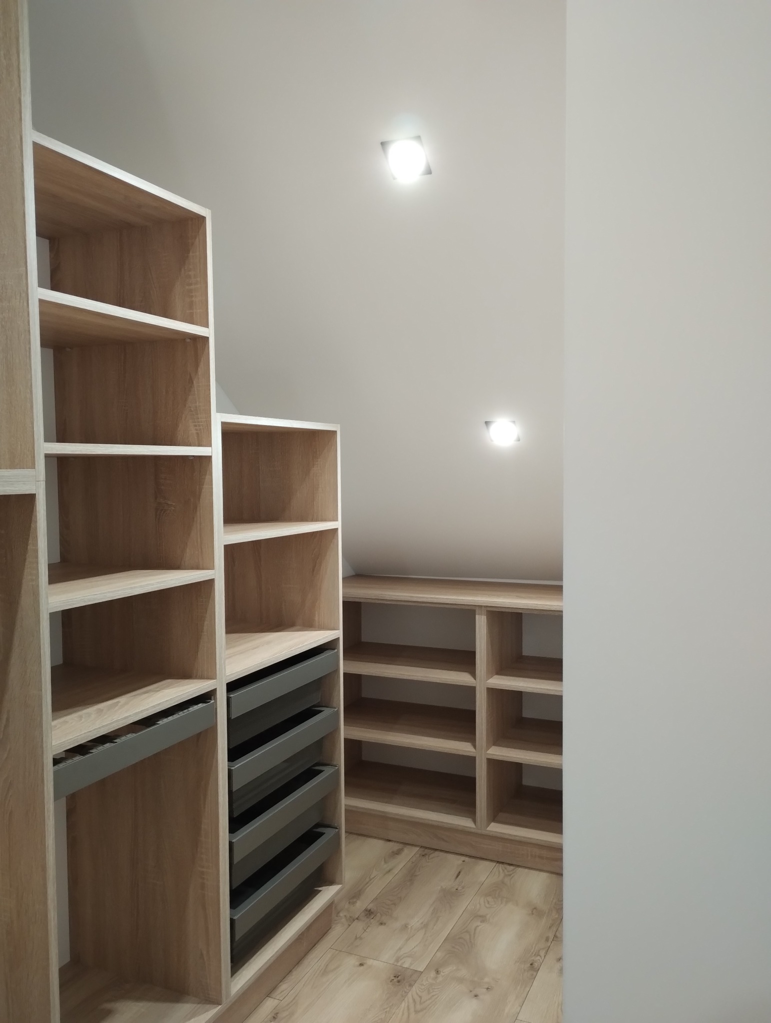 Jasna garderoba z półkami i szufladami w zabudowie pod skośnym sufitem, oświetlona punktowymi lampami.