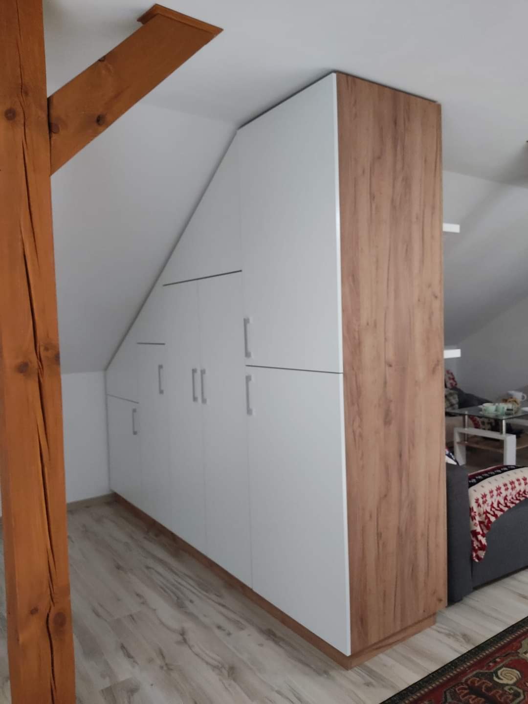 Biała szafa wnękowa na poddaszu w Radoszycach, z drewnianymi elementami wykończeniowymi, dopasowana do skosu dachu, widoczna drewniana belka konstrukcyjna.