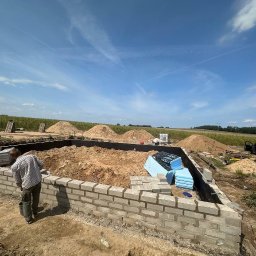 Mr Gruzzly Ihor Papenko - Budowa fundamentów domu z bloczków betonowych; widoczne ułożone bloczki, wykop, hałdy ziemi, materiały izolacyjne oraz dwóch murarzy przy pracy.