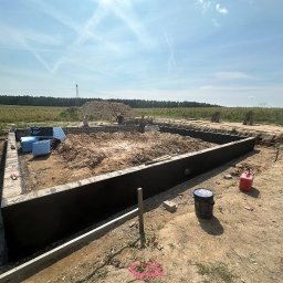 Mr Gruzzly Ihor Papenko - Wykop pod fundamenty z izolacją pionową, częściowo wymurowane ściany fundamentowe z bloczków, składowany styropian do izolacji termicznej, widoczni pracownicy budowlani.