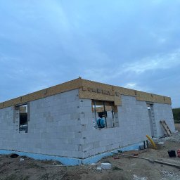 Mr Gruzzly Ihor Papenko - Wznoszenie ścian parterowego domu z betonu komórkowego, widoczna izolacja fundamentów i drewniana konstrukcja wieńcząca ściany, w tle słupy wysokiego napięcia.