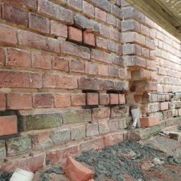 Mr Gruzzly Ihor Papenko - Fragment murowanej ściany z czerwonej cegły w trakcie naprawy, widoczne ubytki i nowo wstawione cegły, pracownik w odzieży roboczej robi zdjęcie telefonem, rusztowanie w tle.