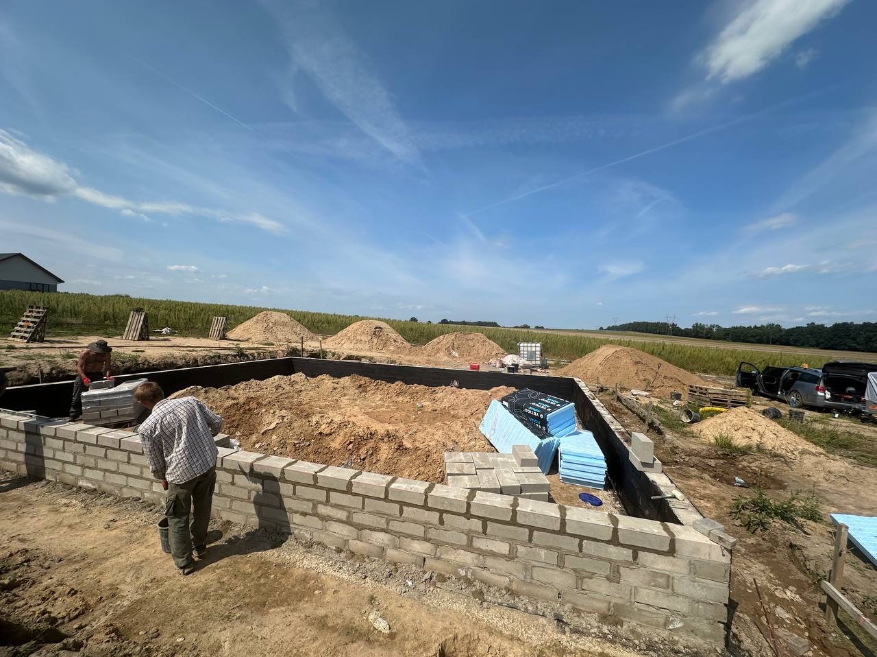 Budowa fundamentów domu z bloczków betonowych; widoczne ułożone bloczki, wykop, hałdy ziemi, materiały izolacyjne oraz dwóch murarzy przy pracy.