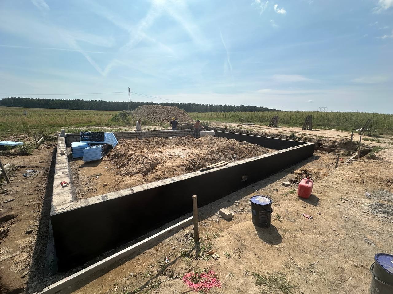 Wykop pod fundamenty z izolacją pionową, częściowo wymurowane ściany fundamentowe z bloczków, składowany styropian do izolacji termicznej, widoczni pracownicy budowlani.