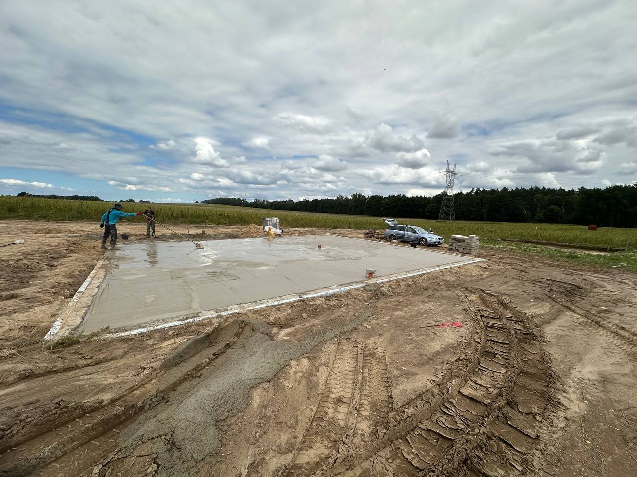 Wylewka betonowa fundamentów domu jednorodzinnego w trakcie prac wykończeniowych, dwóch pracowników wygładza powierzchnię betonu, widoczne ślady opon na błotnistym terenie wokół, w tle samochód...