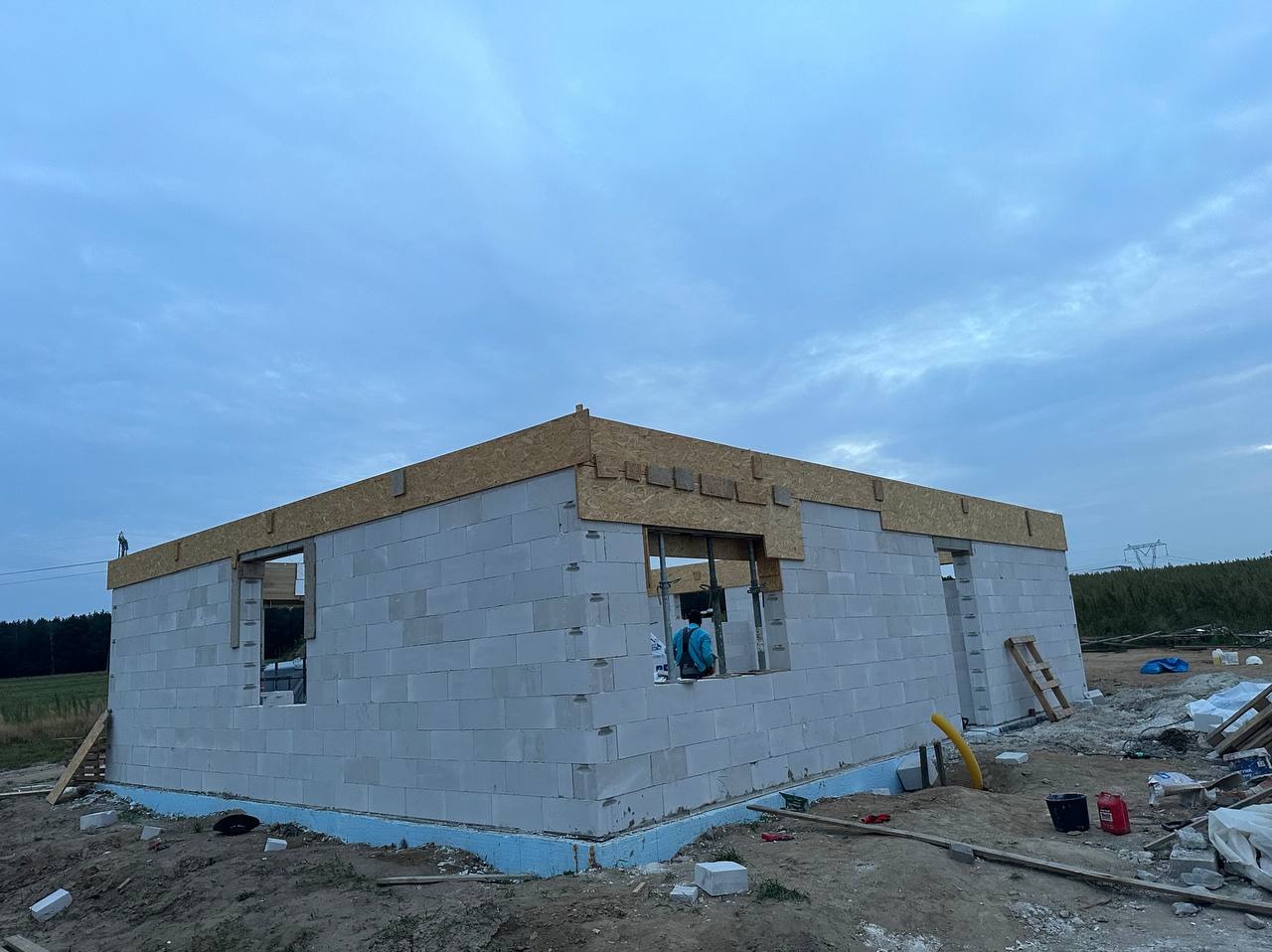 Wznoszenie ścian parterowego domu z betonu komórkowego, widoczna izolacja fundamentów i drewniana konstrukcja wieńcząca ściany, w tle słupy wysokiego napięcia.