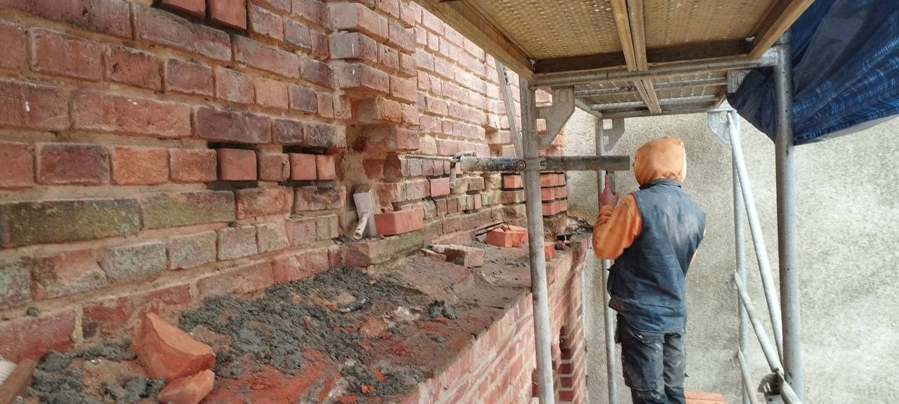 Murarz na rusztowaniu w trakcie naprawy uszkodzonej ściany z czerwonej cegły. Widoczne ubytki, świeża zaprawa i narzędzia murarskie.