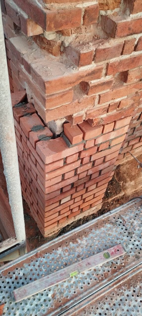 Fragment muru z czerwonej cegły w trakcie wznoszenia, widoczne nierówności i zaprawa, poziomica leży na metalowym rusztowaniu.