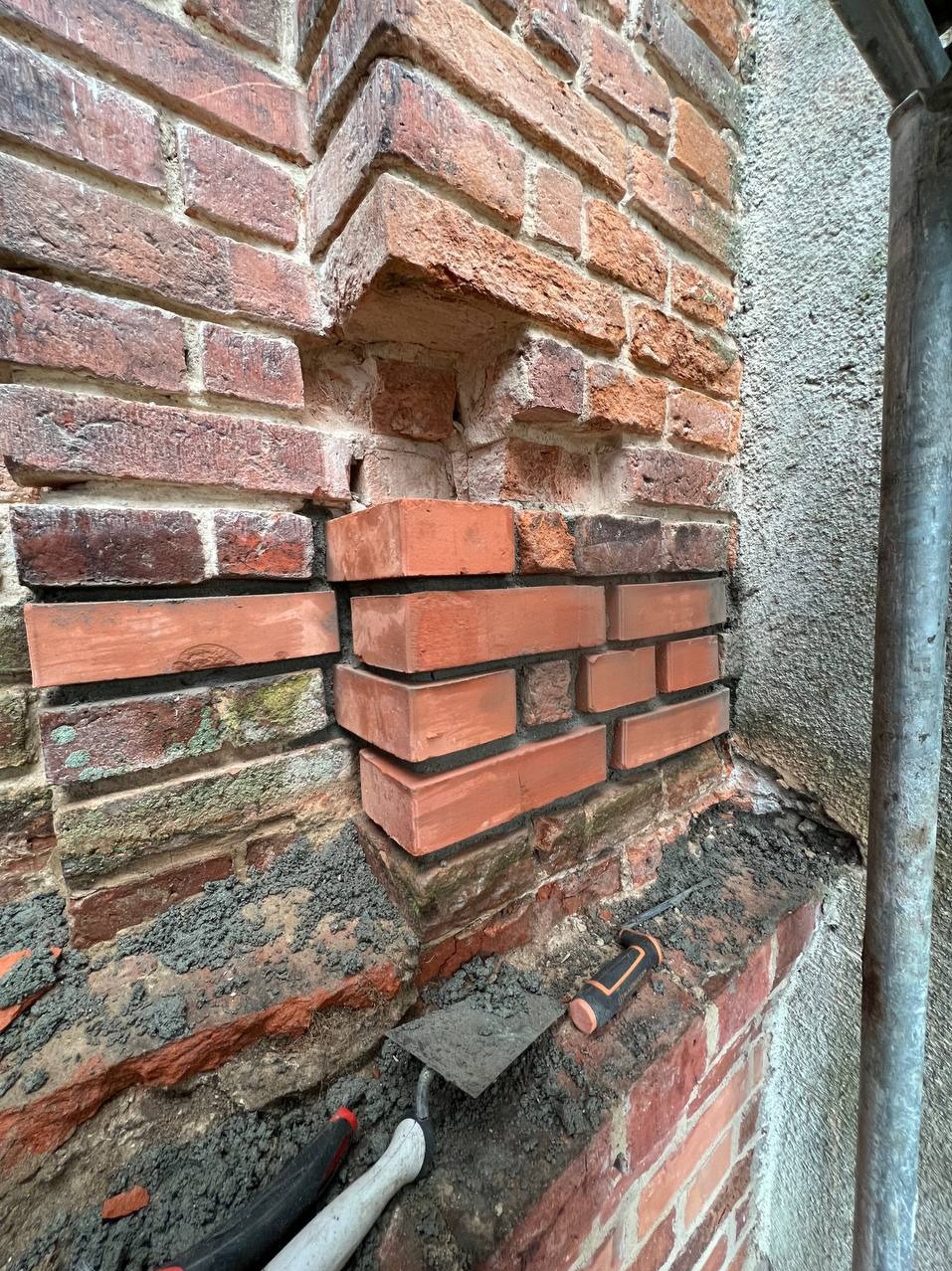 Fragment murowanej ściany z czerwonej cegły w trakcie naprawy, widoczne nowe cegły w miejscu ubytku, kielnia murarska i rusztowanie w tle.