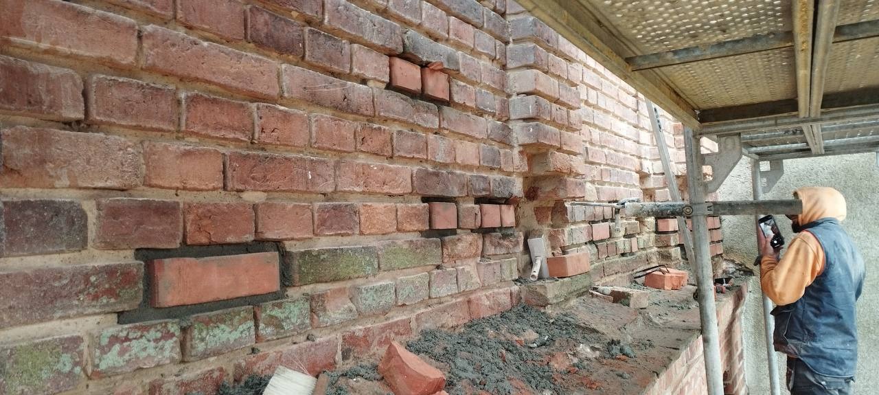 Fragment murowanej ściany z czerwonej cegły w trakcie naprawy, widoczne ubytki i nowo wstawione cegły, pracownik w odzieży roboczej robi zdjęcie telefonem, rusztowanie w tle.