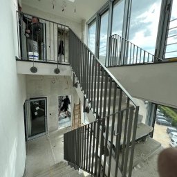 NEUVARTSTAHL SPÓŁKA Z OGRANICZONĄ ODPOWIEDZIALNOŚCIĄ - Wnętrze budynku w trakcie wykańczania z widocznymi schodami z szarą, pionową balustradą, oknami od podłogi do sufitu i osobami pracującymi w tle.