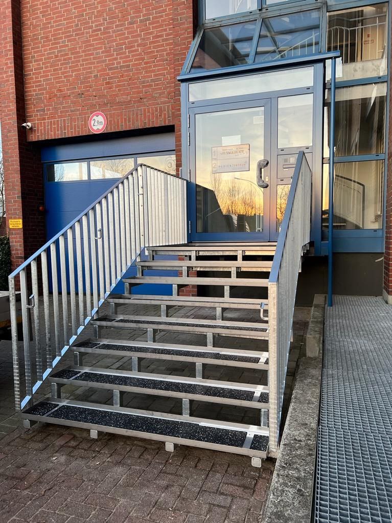 Zewnętrzne schody z metalową konstrukcją i poręczami, prowadzące do przeszklonego wejścia budynku z cegły.