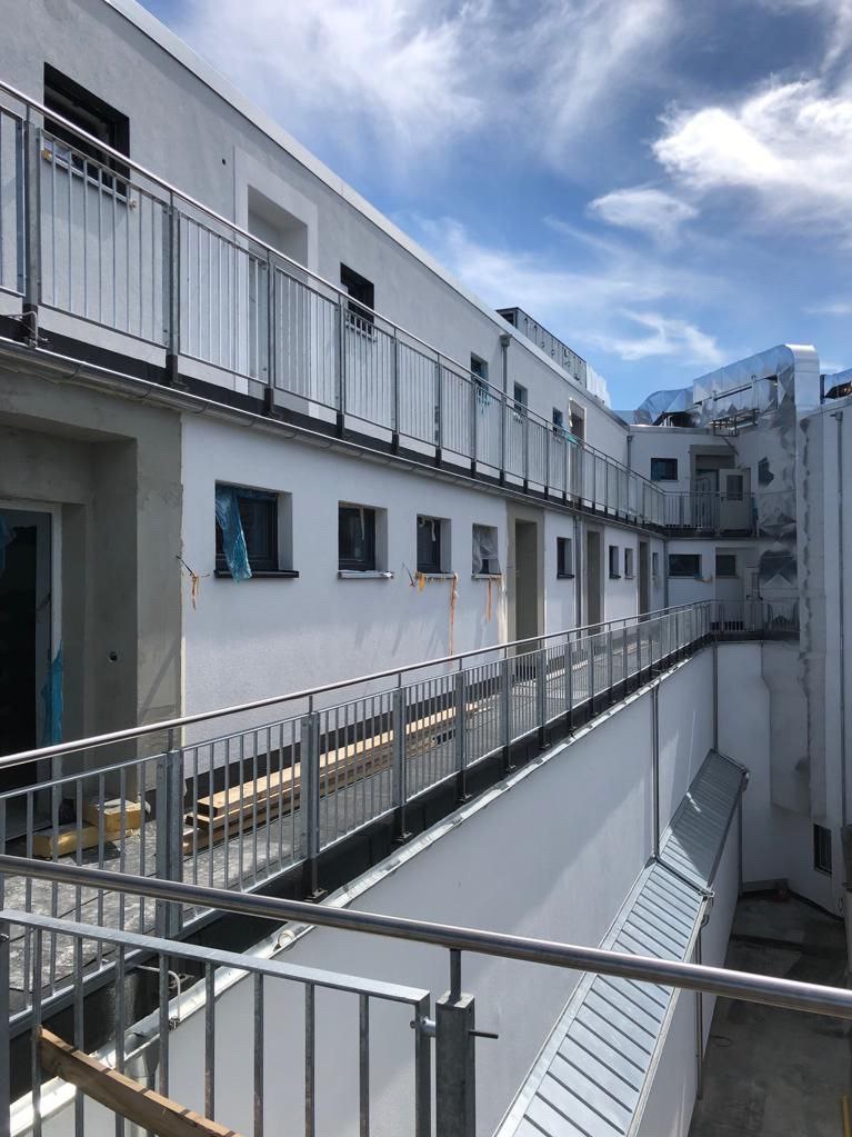 Nowoczesny budynek z balkonami zabezpieczonymi metalowymi balustradami, widoczne elementy wykończeniowe i instalacje wentylacyjne na dachu.