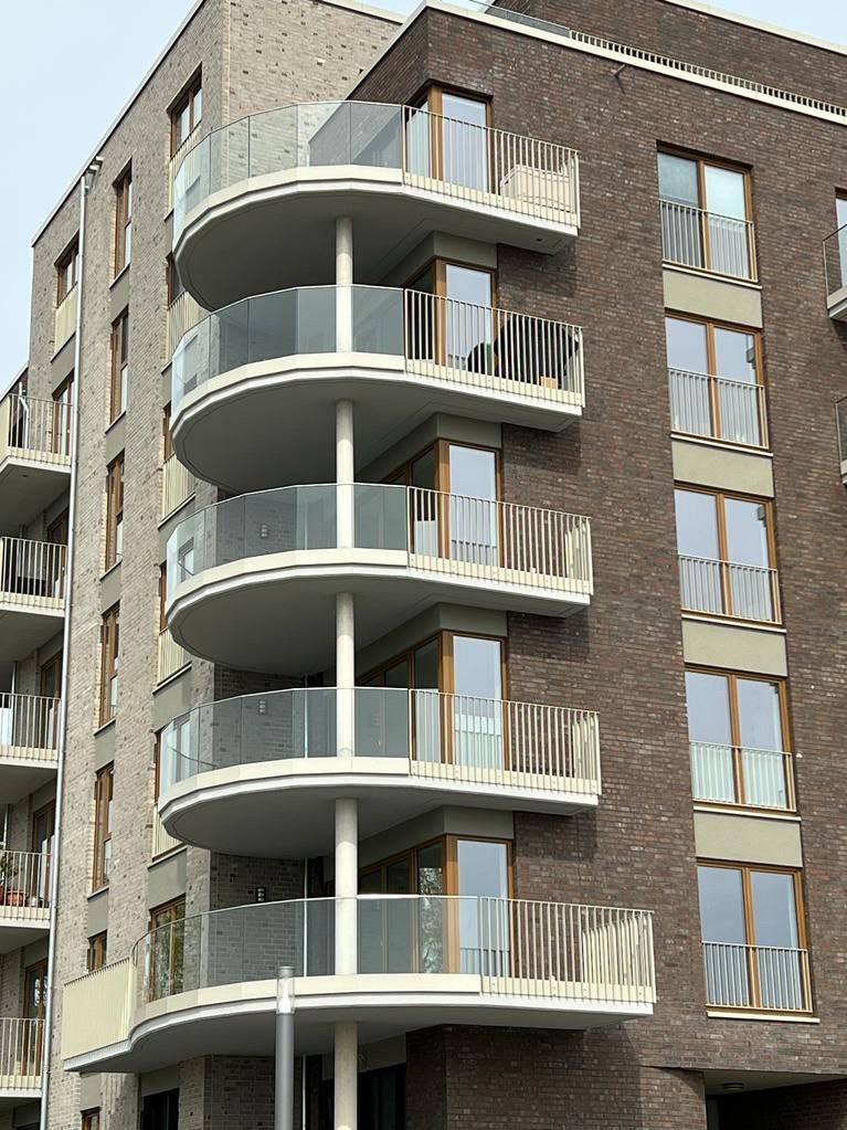 Nowoczesny budynek z zaokrąglonymi balkonami z balustradami ze szkła i metalu, widoczne ceglane elewacje w odcieniach szarości i brązu.
