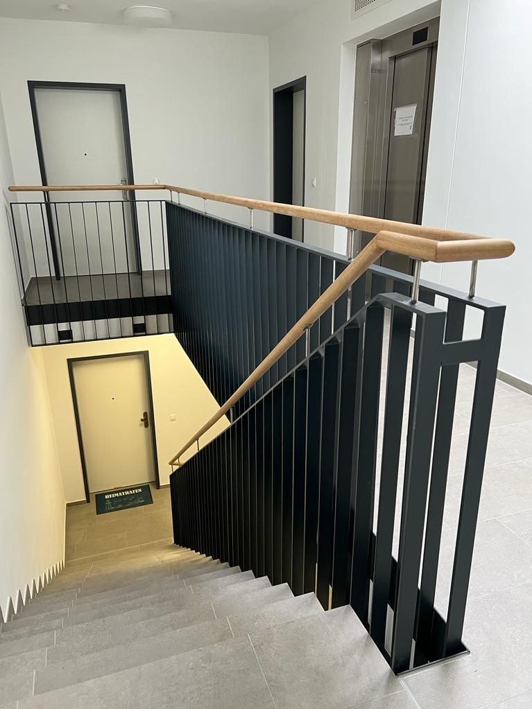 Schody z szarymi stopniami i czarną, metalową balustradą z drewnianą poręczą, widok z góry na klatkę schodową w budynku mieszkalnym z drzwiami i windą w tle.