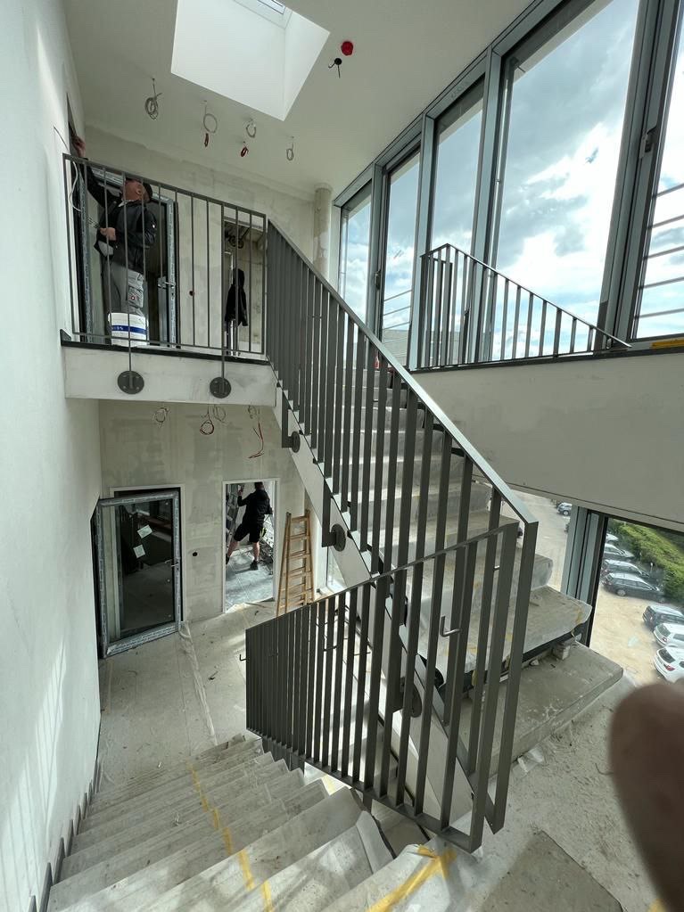 Wnętrze budynku w trakcie wykańczania z widocznymi schodami z szarą, pionową balustradą, oknami od podłogi do sufitu i osobami pracującymi w tle.