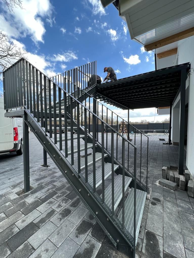 Montaż zewnętrznych schodów z metalu prowadzących do zadaszonej pergoli, widoczne dwie osoby pracujące na górze, obok zaparkowany biały samochód dostawczy.