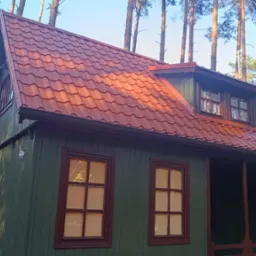 Zielony drewniany domek z czerwoną, metalową dachówką w lesie, widoczne okna z brązowymi ramami i rynny.
