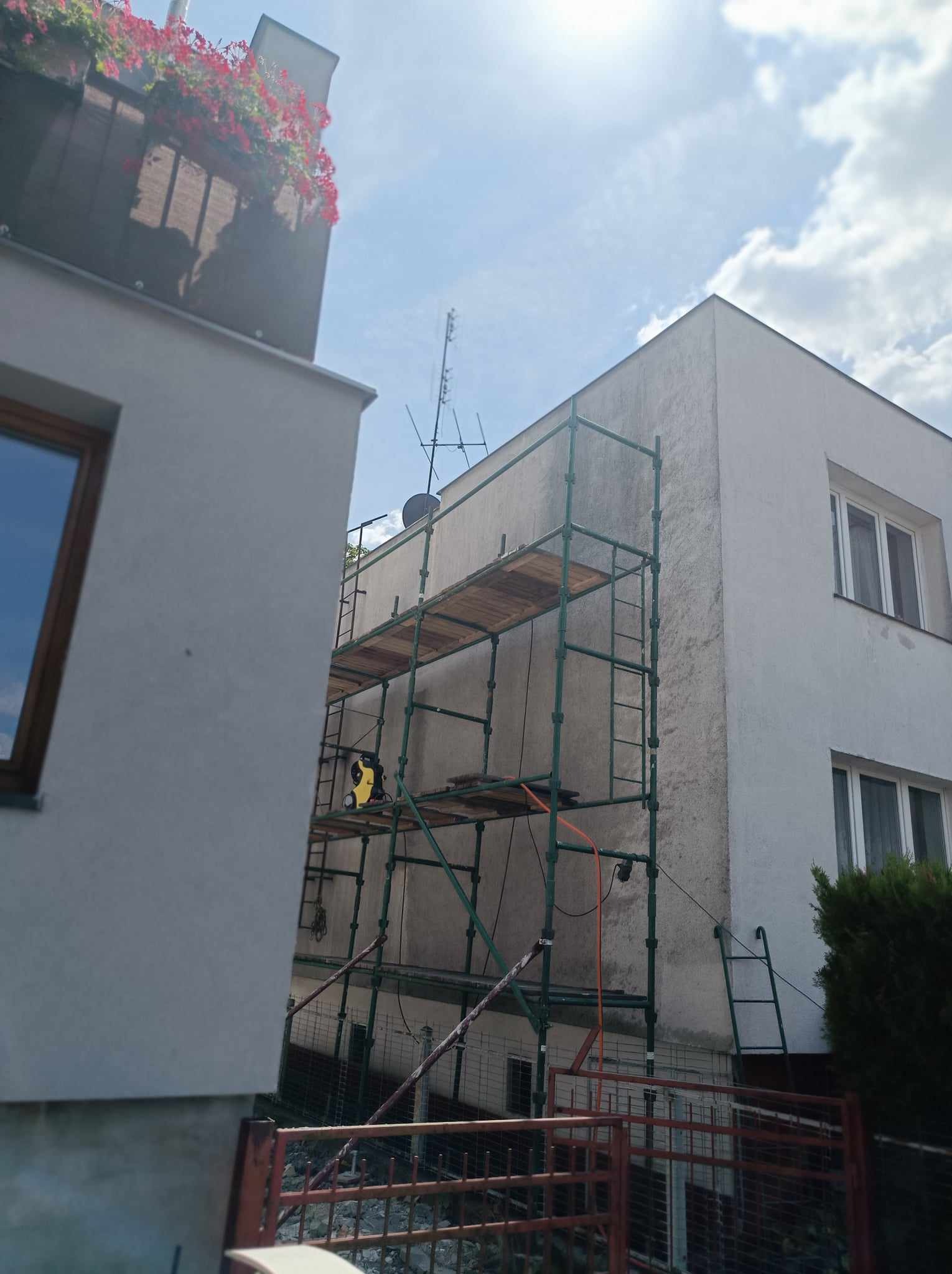 Budynek dwukondygnacyjny w trakcie renowacji elewacji, widoczne rusztowanie, drabina oparta o ścianę i żółta myjka ciśnieniowa na platformie rusztowania.