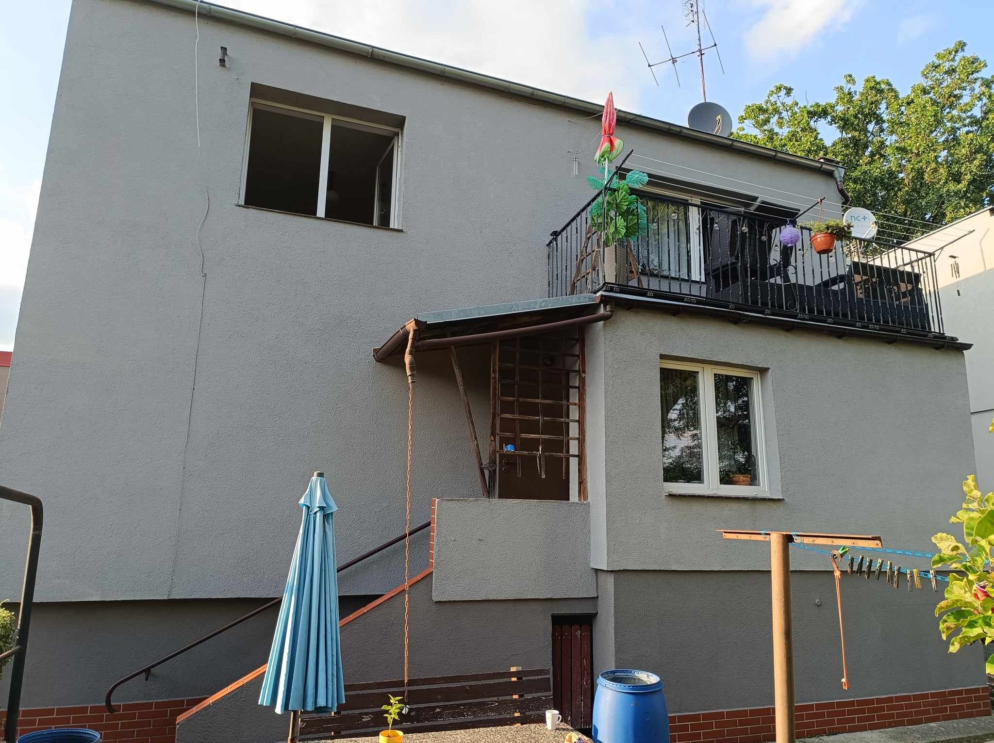 Szary dom jednorodzinny w Sulechowie z balkonem i widoczną elewacją, na której widać ślady upływu czasu. Na balkonie rośliny i przedmioty codziennego użytku. Dolna część elewacji pomalowana...