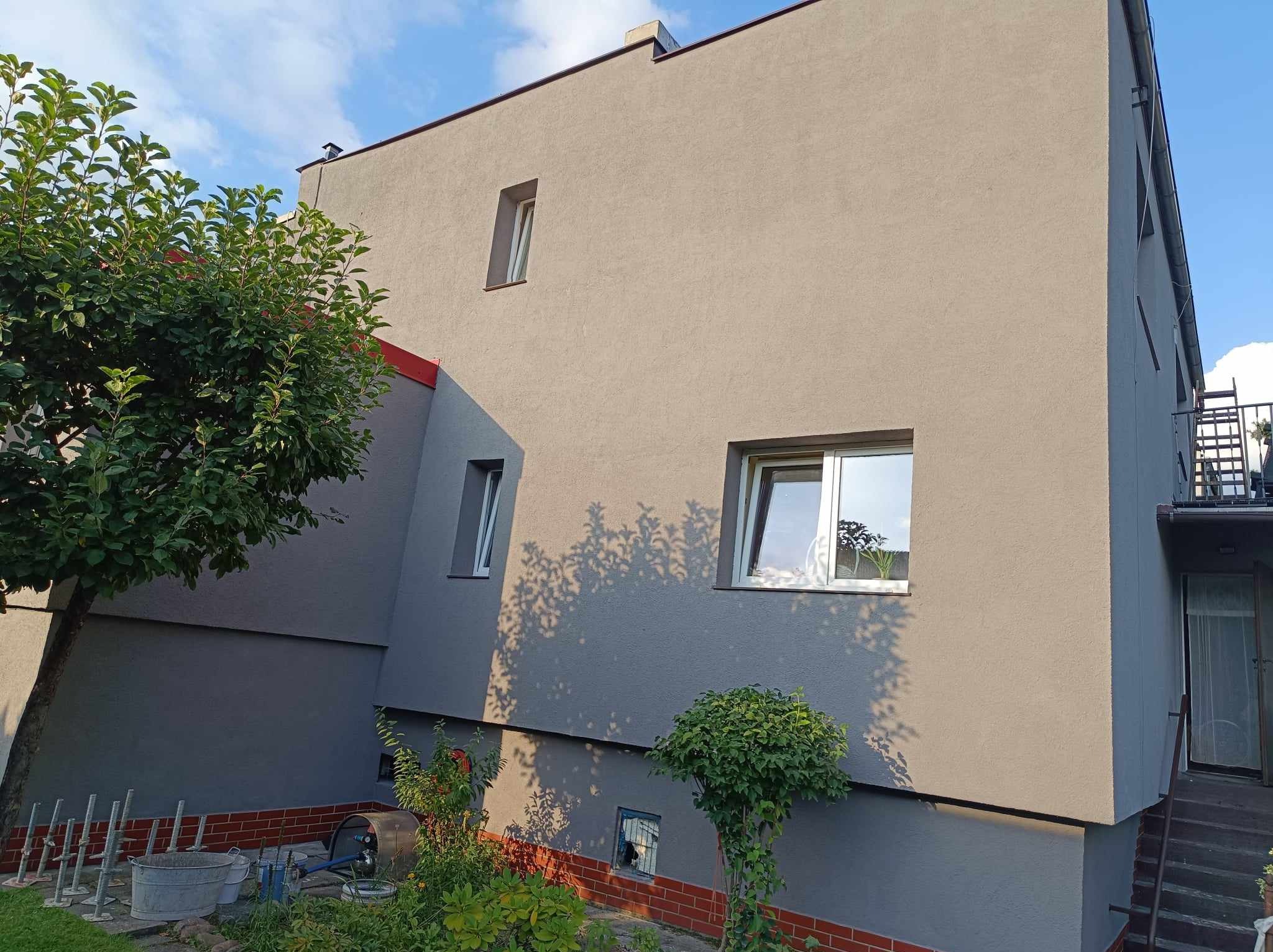 Ocieplony i pomalowany budynek mieszkalny z widocznymi oknami, fragmentem dachu, zielenią i narzędziami budowlanymi w otoczeniu.