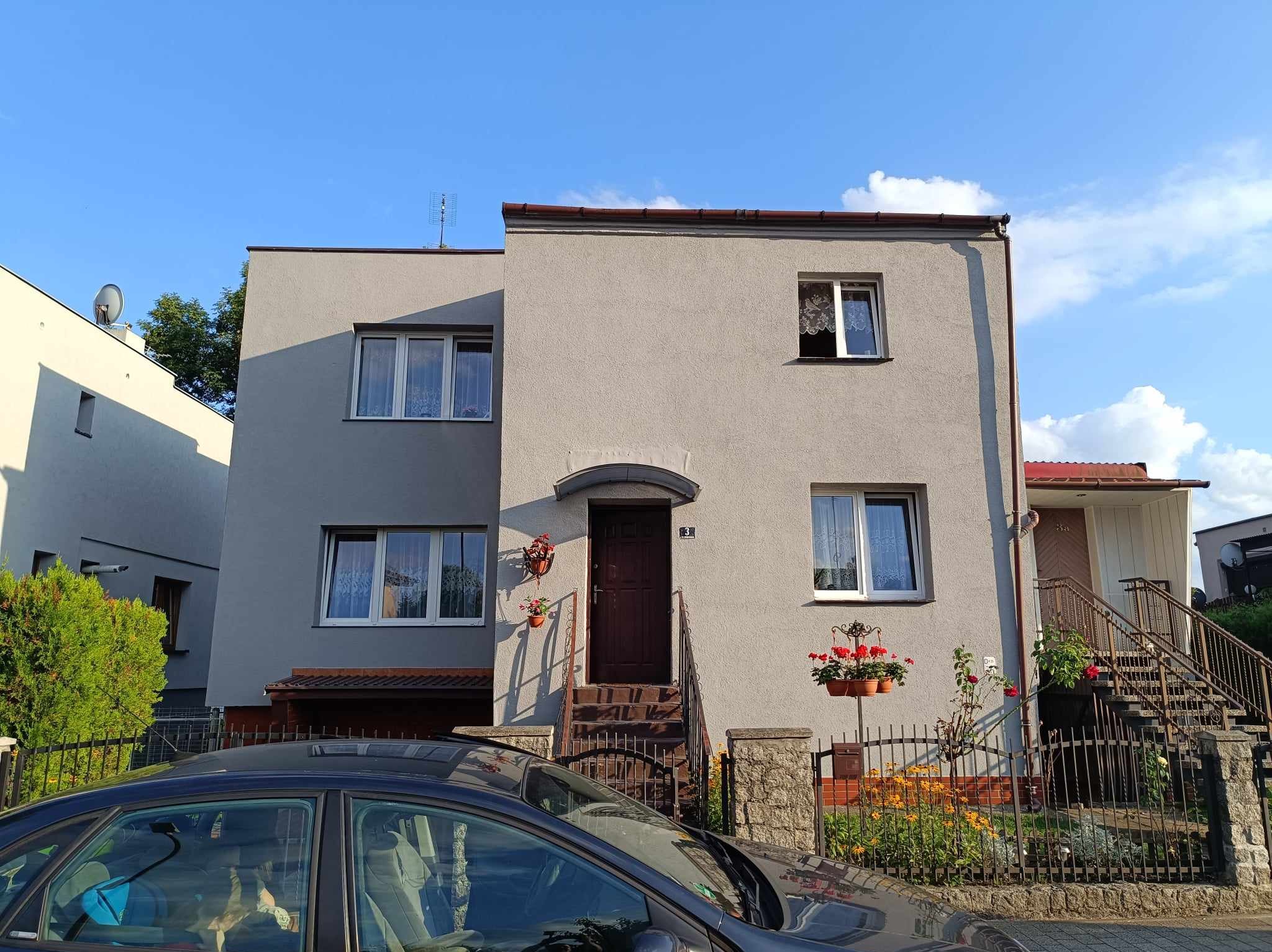 Dwu-kondygnacyjny dom z szarą elewacją, ciemnymi ramami okiennymi i brązowymi drzwiami wejściowymi, z roślinami w doniczkach na elewacji i przy wejściu, widziany z poziomu ulicy, z częściowo...