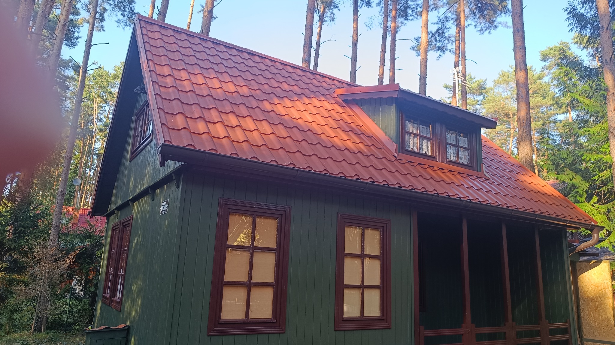 Zielony drewniany domek z czerwoną, metalową dachówką w lesie, widoczne okna z brązowymi ramami i rynny.