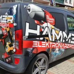 Czarny samochód dostawczy z grafiką narzędzi i napisem HAMMER ELEKTRONARZĘDZIA, zaparkowany na brukowanej powierzchni.