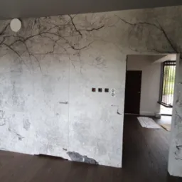 Wnętrze z fototapetą imitującą spękaną ścianę i motyw drzew, ukryte drzwi z minimalistyczną klamką oraz widoczne gniazdka elektryczne i włączniki.