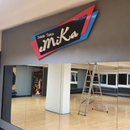SD Projekt Marcin Sanduła - Wnętrze sali tanecznej z dużym, podświetlonym logo 'Szkoła Tańca eMiKa' na ścianie, drabina oparta o lustro, parkiet, okna i plakat w tle.