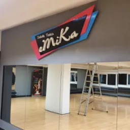 Wnętrze sali tanecznej z dużym, podświetlonym logo 'Szkoła Tańca eMiKa' na ścianie, drabina oparta o lustro, parkiet, okna i plakat w tle.
