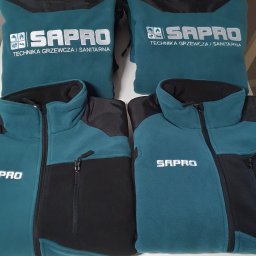SD Projekt Marcin Sanduła - Dwie turkusowo-czarne bluzy polarowe i dwa worki ze srebrnym napisem 'SAPRO Technika Grzewcza i Sanitarna' leżące na białym tle.