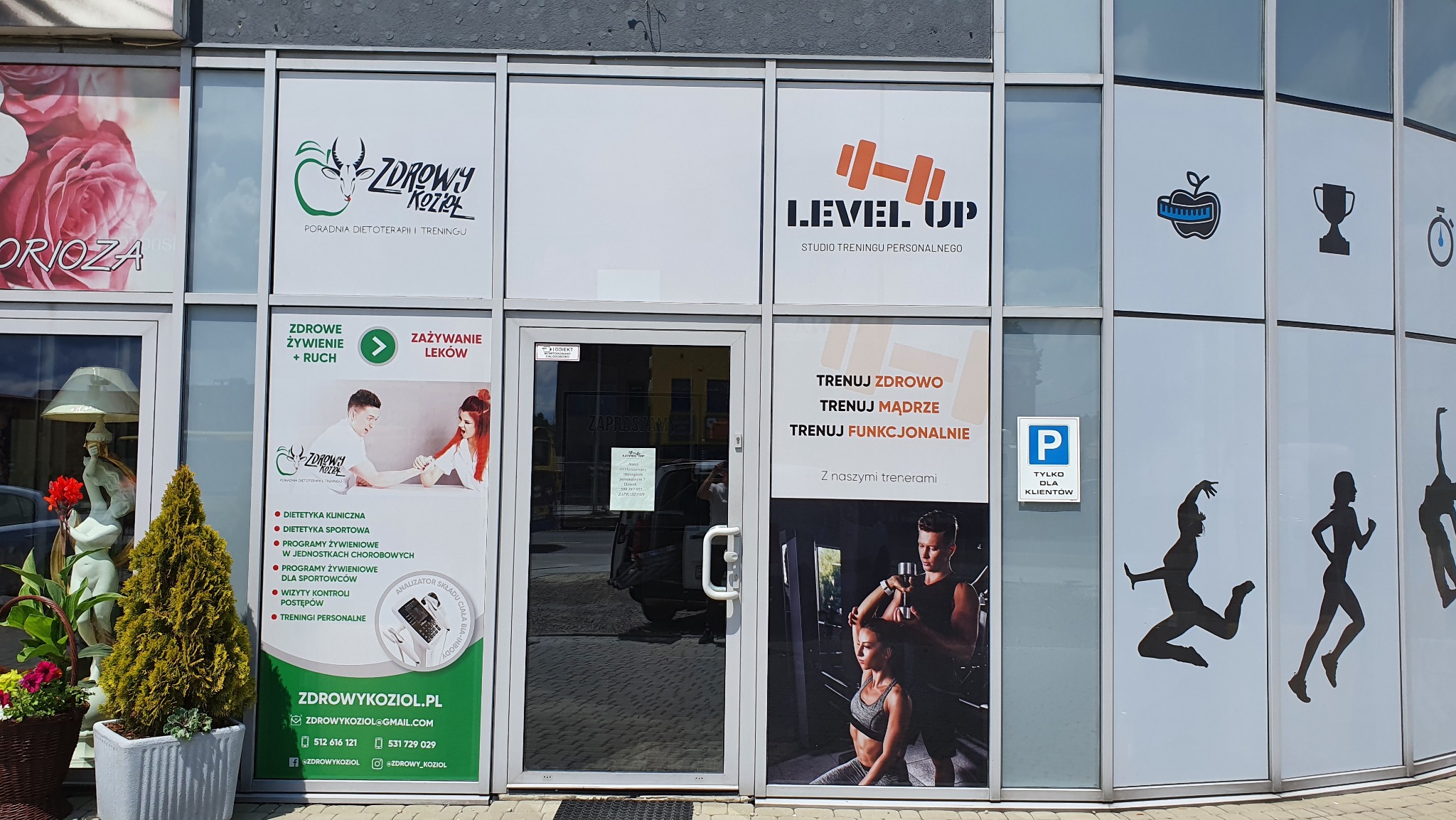 Fasada budynku z witrynami reklamującymi poradnię dietoterapii i studio treningu personalnego, z widocznymi logotypami, informacjami o usługach i sylwetkami ćwiczących.
