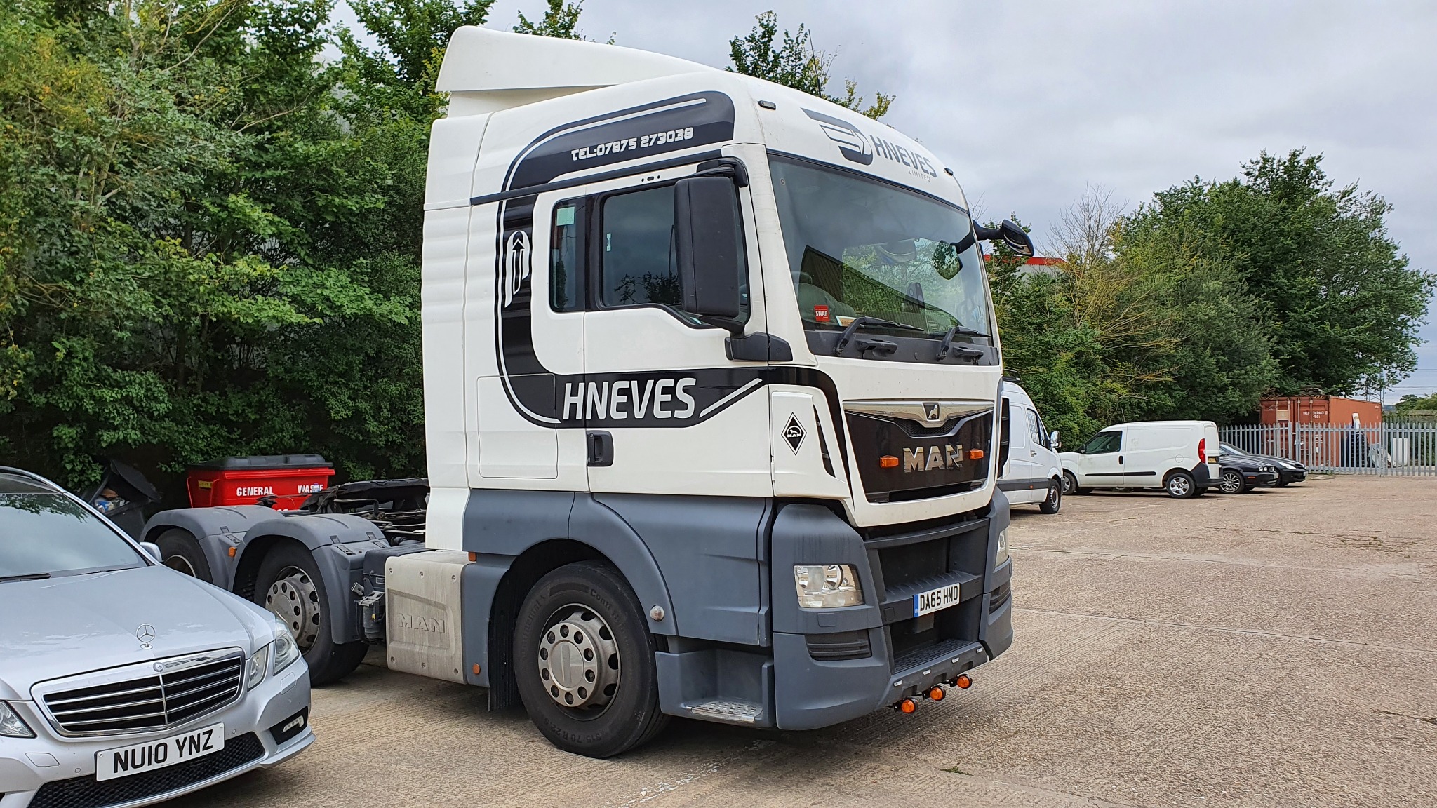 Ciągnik siodłowy MAN z naniesionym logo i nazwą firmy HNEVES w czarno-białej kolorystyce, zaparkowany na placu z widocznym srebrnym samochodem osobowym i innymi pojazdami w tle.