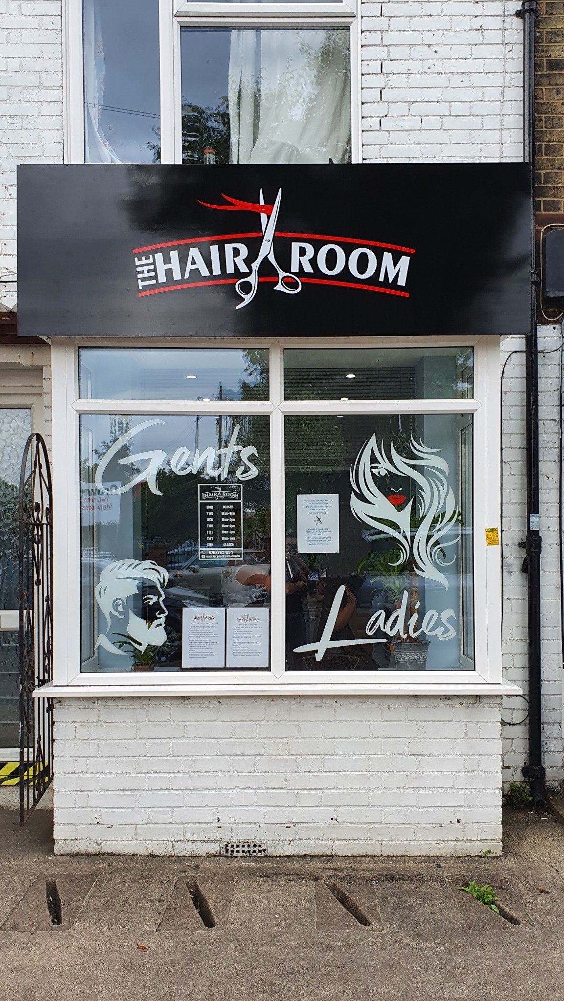 Fasada salonu fryzjerskiego z czarnym szyldem z napisem 'The Hair Room' i grafiką nożyczek. Na oknach naklejki z napisami 'Gents' i 'Ladies' oraz sylwetkami twarzy.