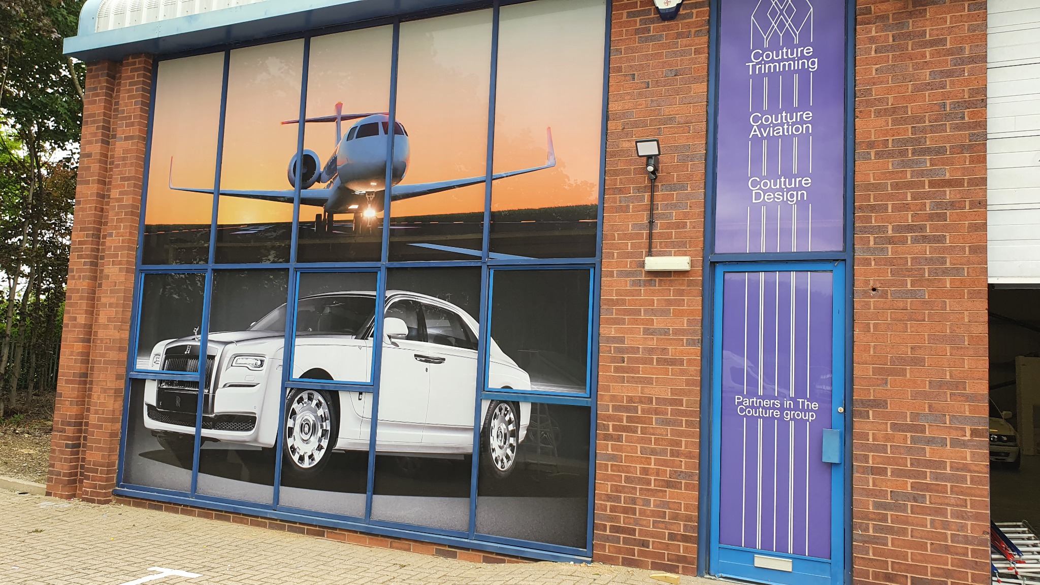 Zewnętrzny widok budynku z dużym oknem oklejonym grafiką przedstawiającą luksusowy samochód i samolot na tle zachodzącego słońca, obok wejście z fioletowym napisem 'Couture Trimming Aviation Design'.