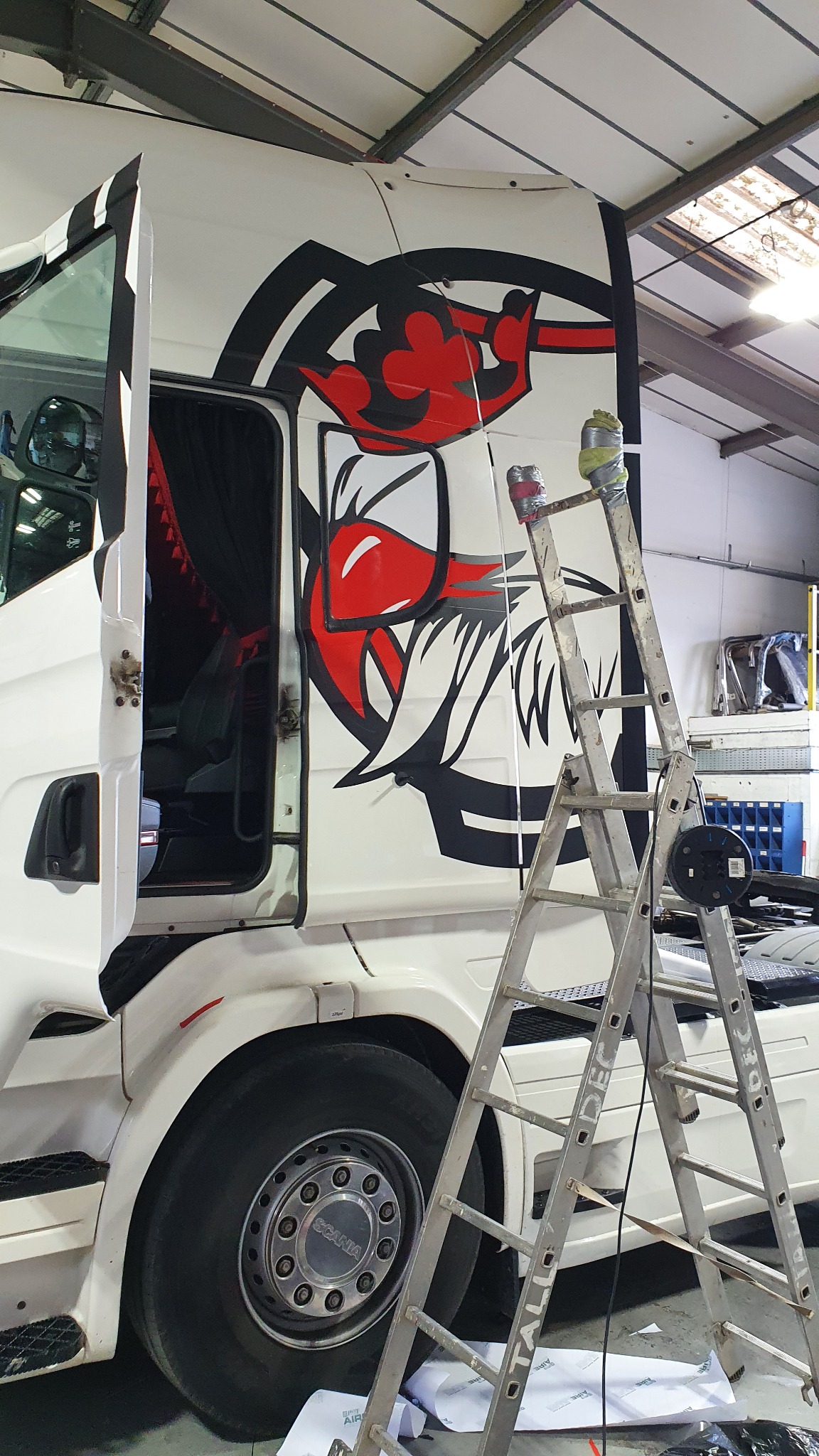Biała ciężarówka z otwartymi drzwiami i naklejoną grafiką w czarno-czerwonej kolorystyce, widoczna aluminiowa drabina z puszkami na górze, w tle warsztat.