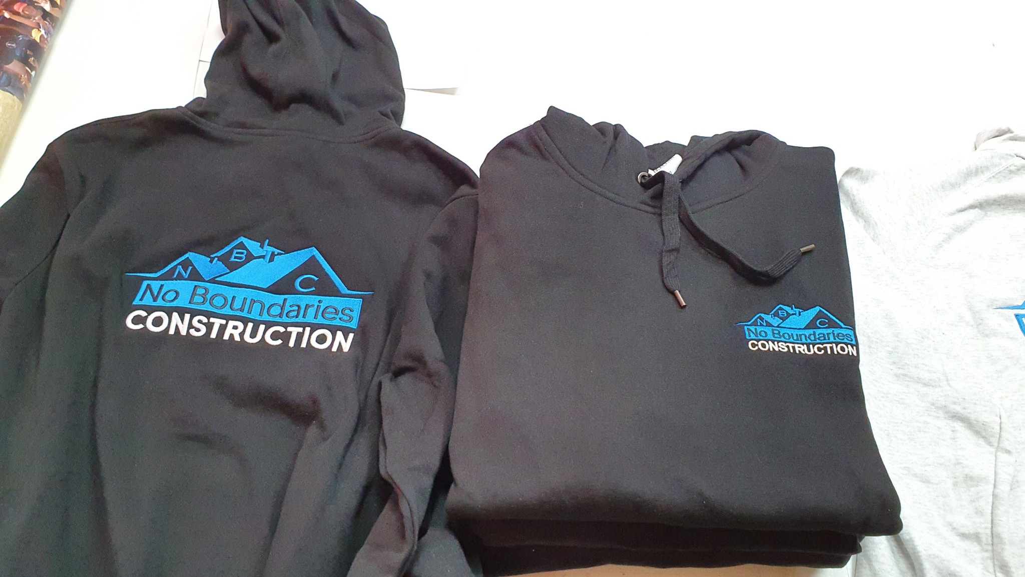 Dwa czarne i jeden szary sweter z kapturem z nadrukowanym niebieskim logo 'No Boundaries Construction'.