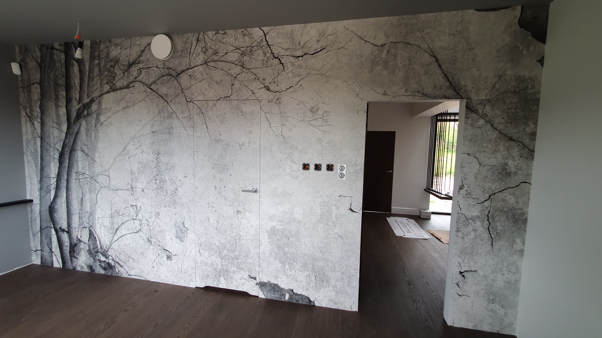 Wnętrze z fototapetą imitującą spękaną ścianę i motyw drzew, ukryte drzwi z minimalistyczną klamką oraz widoczne gniazdka elektryczne i włączniki.