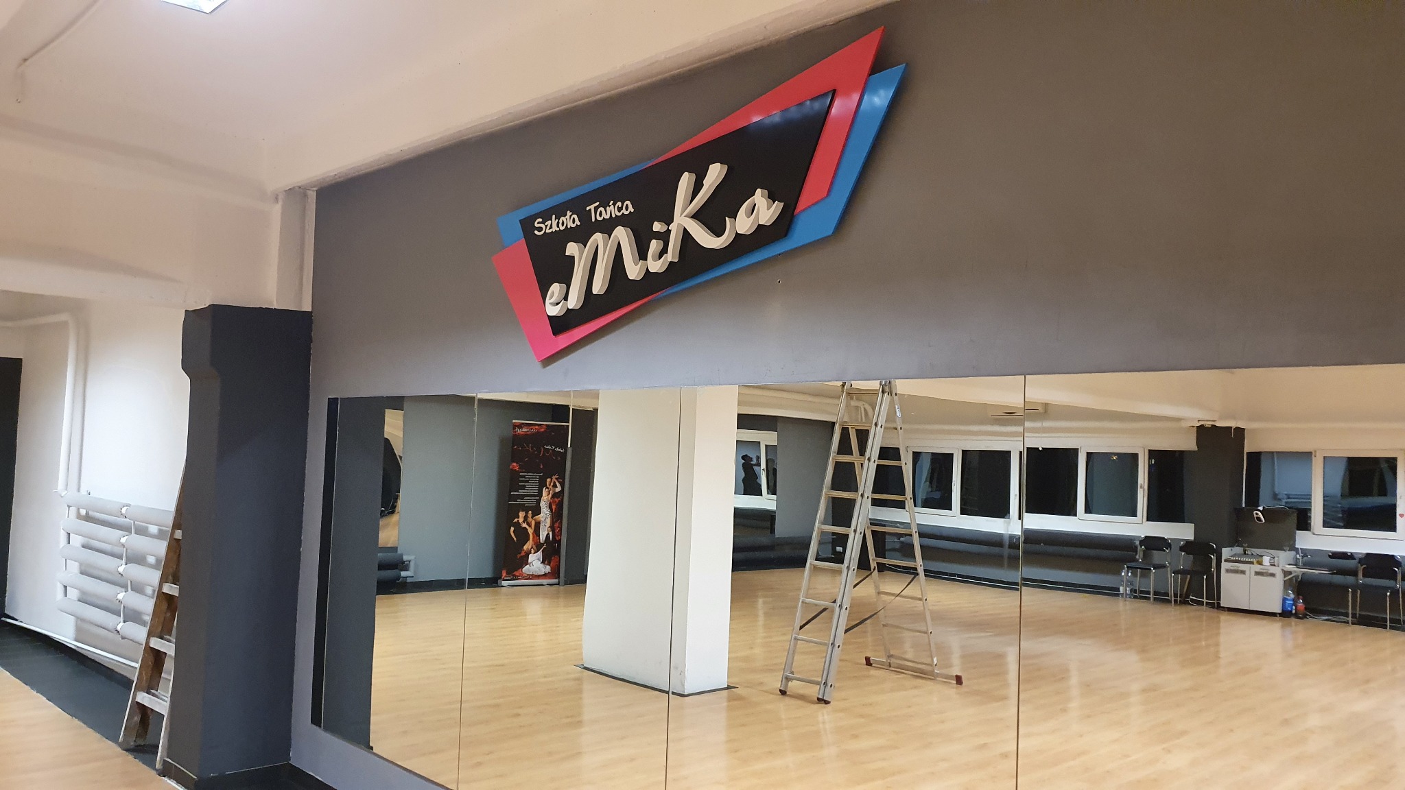 Wnętrze sali tanecznej z dużym, podświetlonym logo 'Szkoła Tańca eMiKa' na ścianie, drabina oparta o lustro, parkiet, okna i plakat w tle.