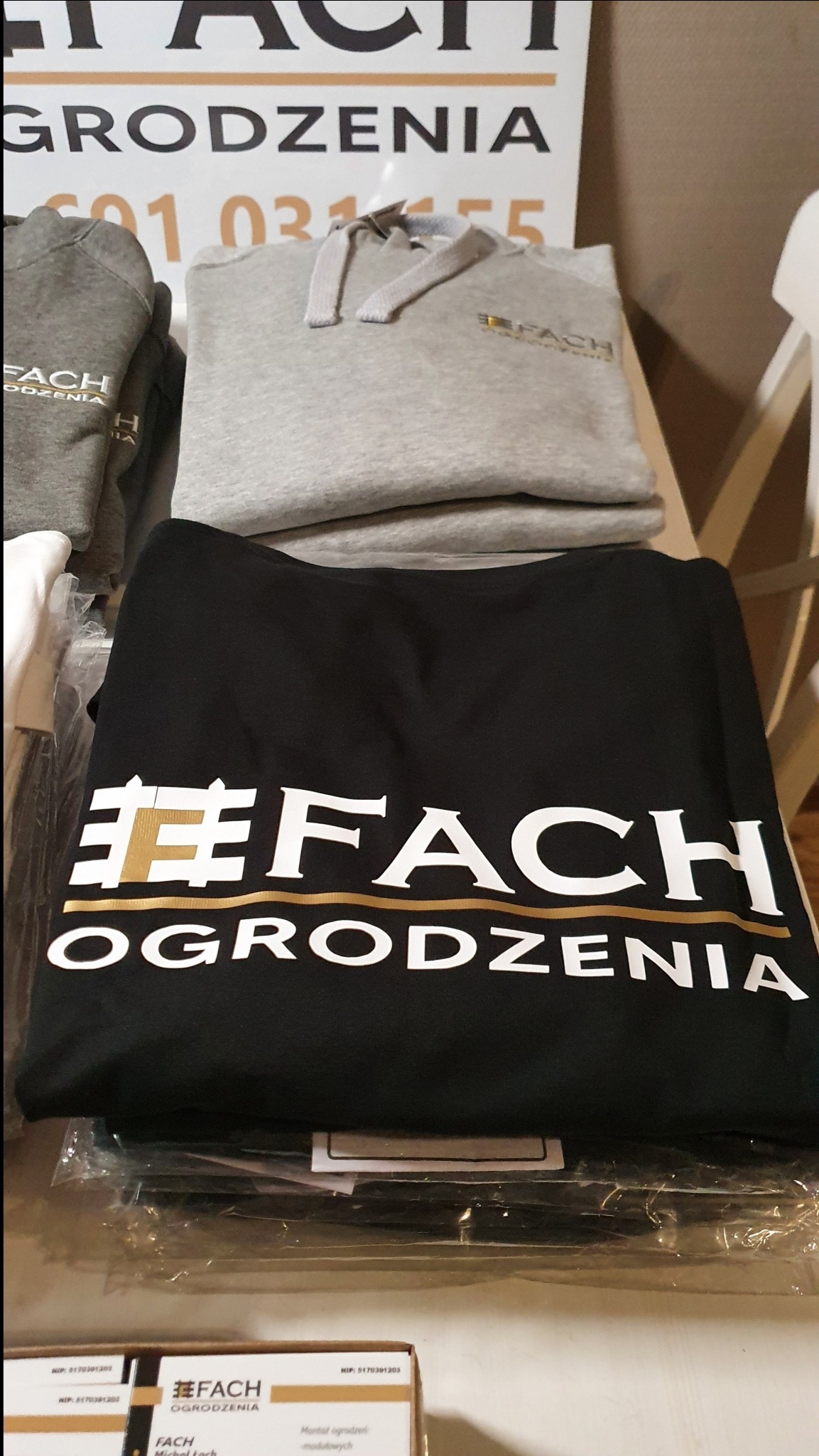 Stos ubrań firmowych, czarna koszulka i szara bluza z kapturem, z białym nadrukiem logo firmy ogrodzeniowej.