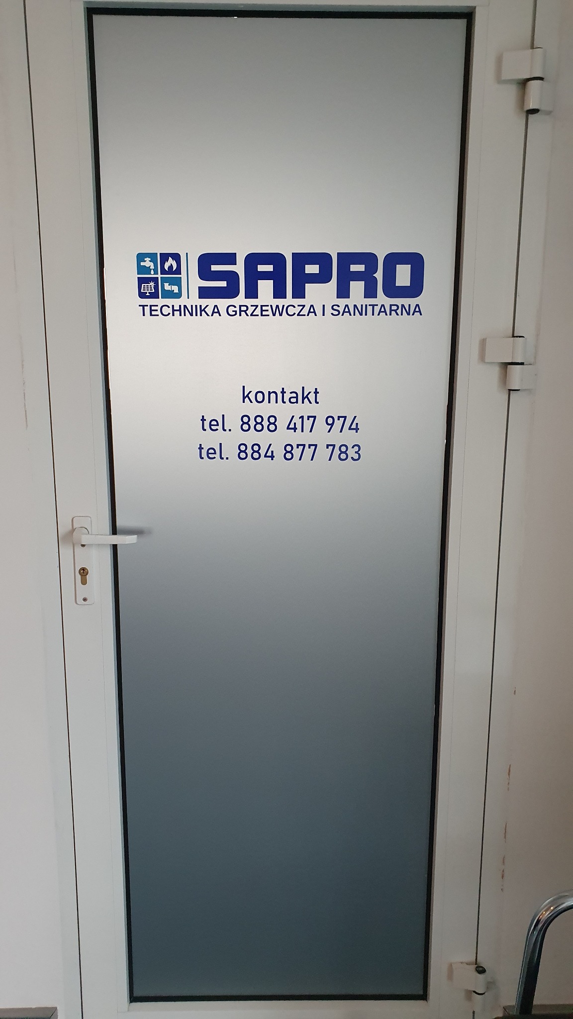 Oklejona folią matową szyba drzwi z logo firmy SAPRO, specjalizującej się w technice grzewczej i sanitarnej, oraz danymi kontaktowymi w kolorze niebieskim.