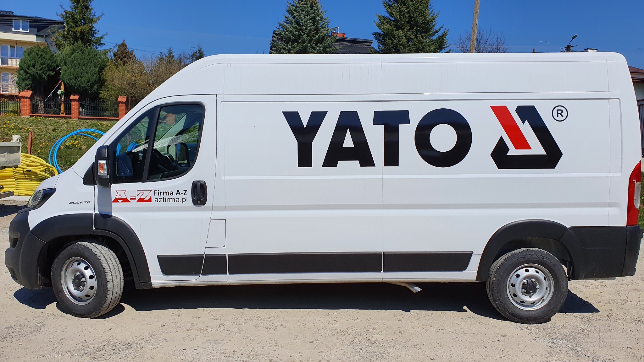 Biały samochód dostawczy Fiat Ducato z czarnym logo YATO i czerwonym akcentem, zaparkowany na żwirowym podłożu, z widocznym logo firmy A-Z na drzwiach kabiny.