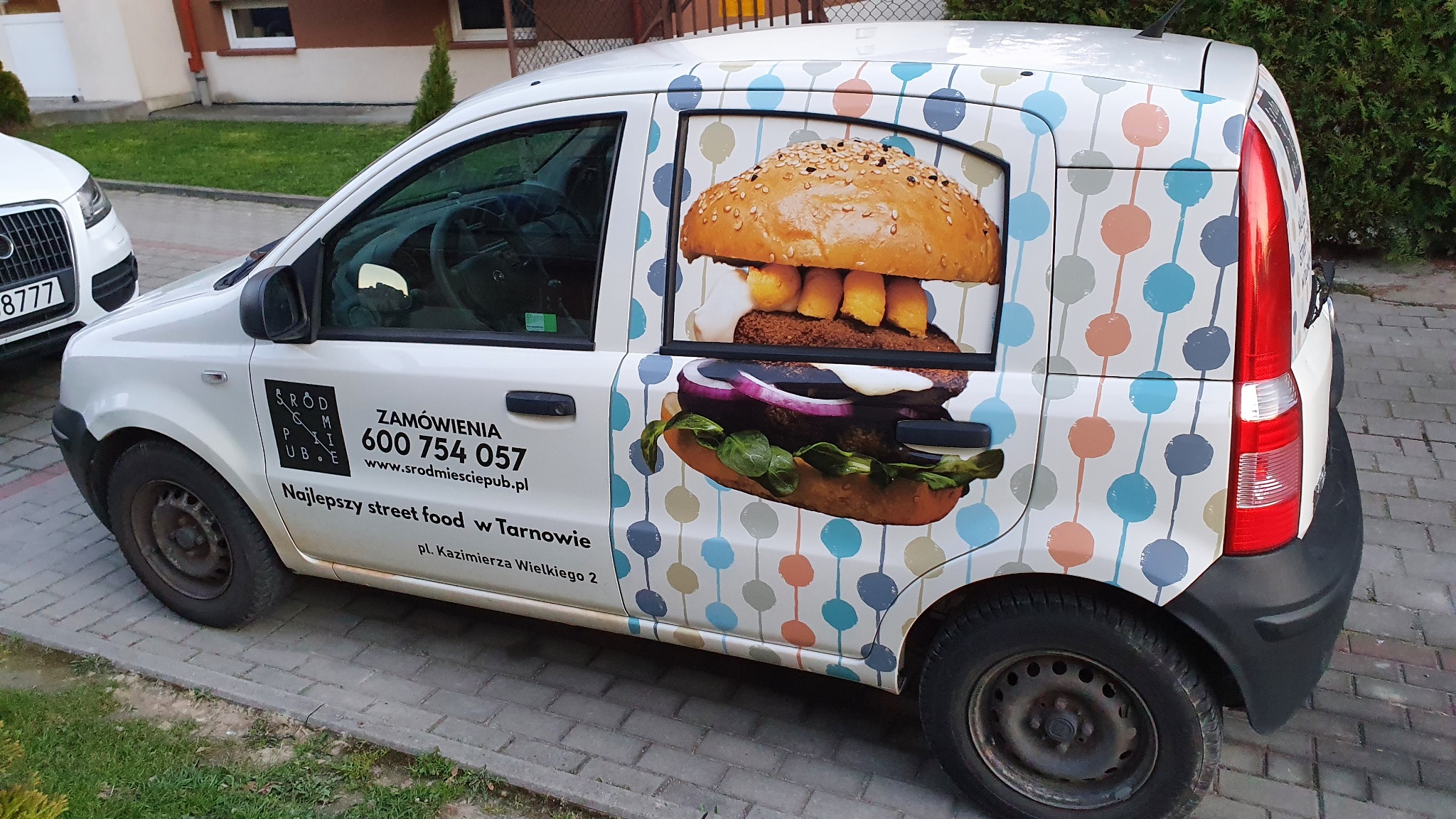 Biały samochód dostawczy z reklamą street foodu: kolorowe kropki i grafika hamburgera na karoserii, numer telefonu i adres strony internetowej na drzwiach.
