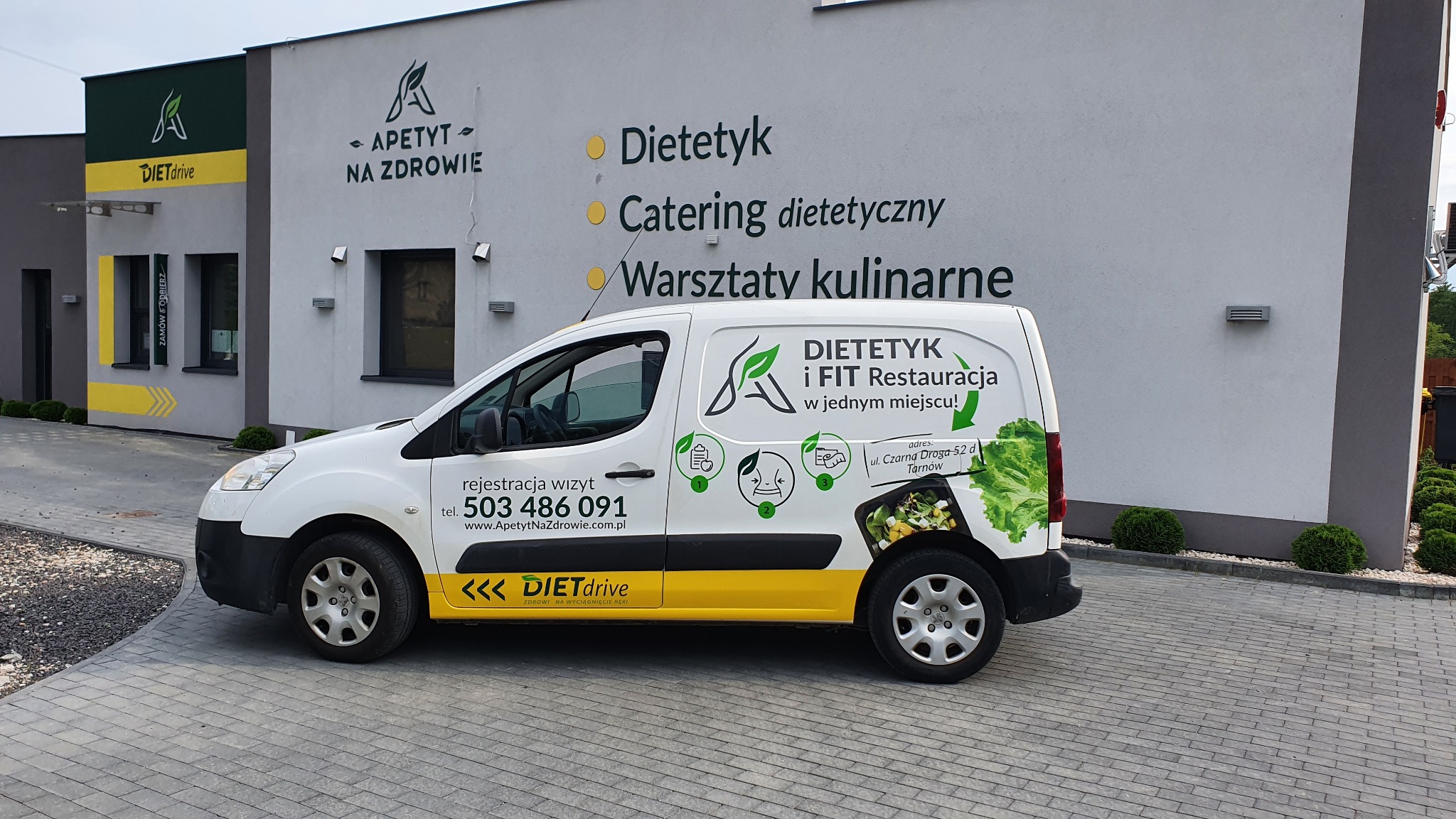 Biała furgonetka z żółtymi akcentami, oklejona grafiką reklamową firmy 'Dietetyk i FIT Restauracja', zaparkowana przed budynkiem z logo 'Apetyt na zdrowie' i informacją o dietetyku, cateringu...