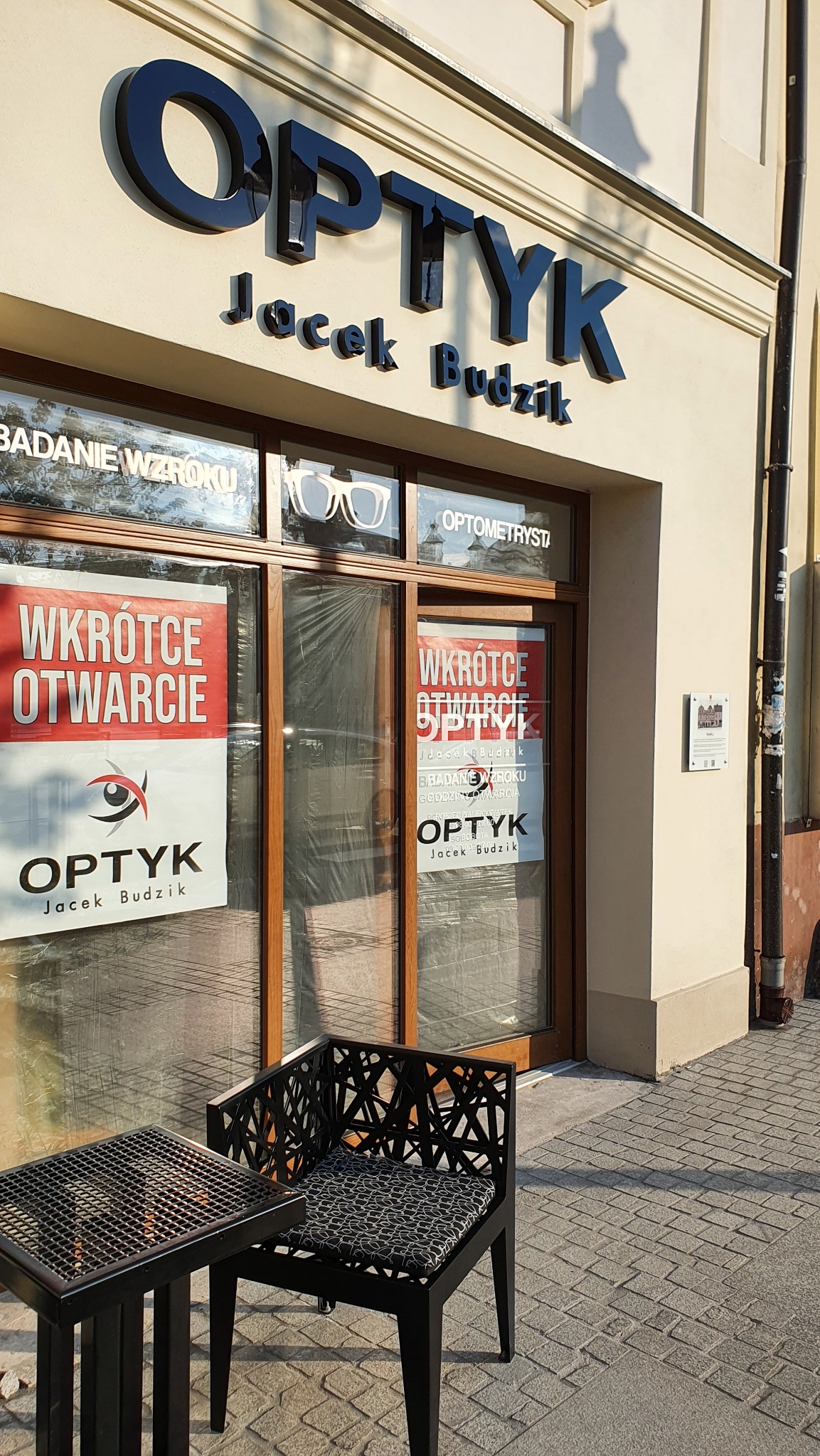 Fasada budynku optyka z szyldem 'OPTYK Jacek Budzik', plakatami 'Wkrótce otwarcie' na witrynie oraz stolikiem i krzesłem przed wejściem.