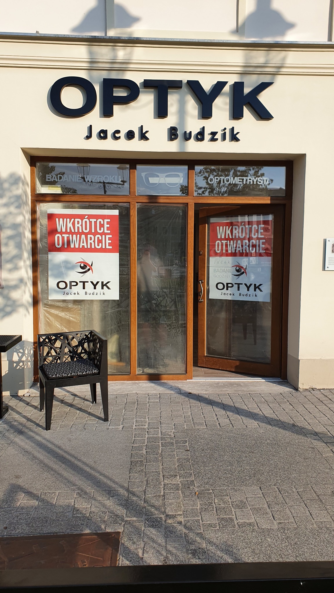 Fasada nowo otwartego salonu optycznego 'Optyk Jacek Budzik' w Tarnowie, z plakatem 'Wkrótce Otwarcie' na witrynie i czarnym krzesłem dla klientów przed wejściem.